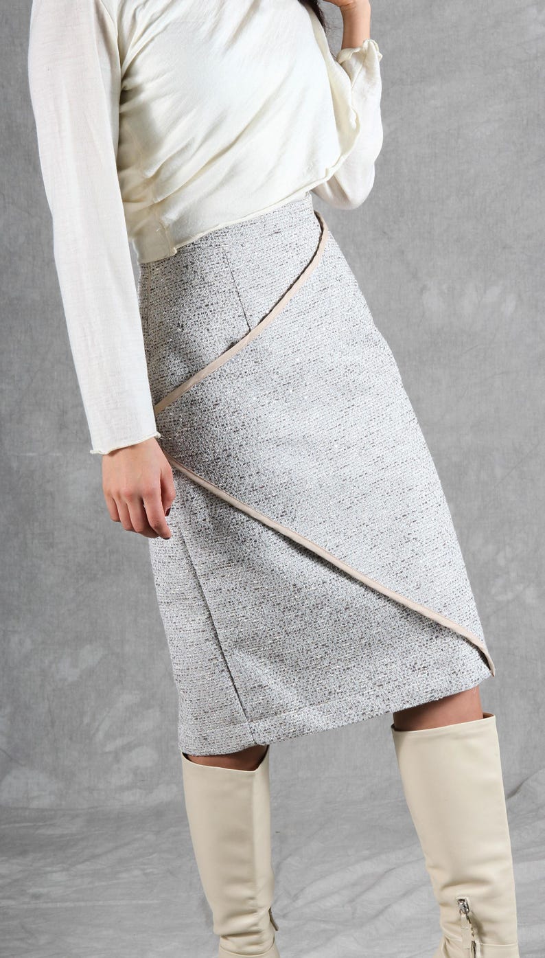 K&ouml;nnte beinhalten: Ein knielanger Rock aus strukturiertem grau-wei&szlig;em Tweed-Stoff. Der Rock verf&uuml;gt &uuml;ber ein Vorderteil in Wickeloptik mit einer diagonalen Naht, die durch eine glatte, hellbeige Borte akzentuiert wird, die von der Taille bis zum Saum verl&auml;uft.