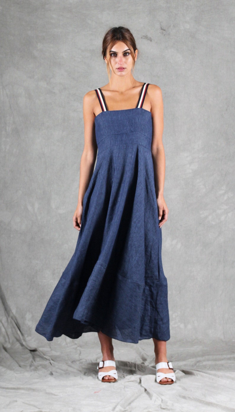 May include: A dark blue midi dress with a square neckline and wide shoulder straps featuring red, white, and blue stripes. The dress has a fitted bodice and a flared, flowing skirt that reaches below the knees, paired with white open-toe sandals.