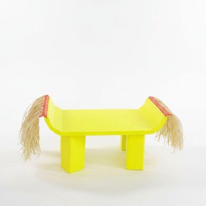 Puede incluir: Un banco de color amarillo brillante con cuatro patas robustas y extremos curvados. Cada extremo presenta un borde rojo con largos flecos fibrosos de color claro que cuelgan de los lados, creando una textura distintiva en este mueble bajo.