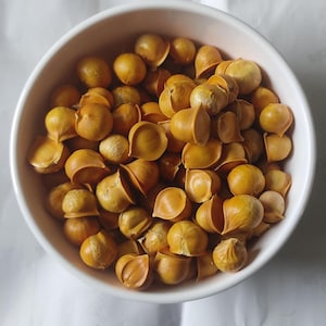 May include: A white bowl filled with numerous golden-brown garlic cloves. The cloves are partially peeled, revealing the inner segments. The image is taken from a top-down perspective, highlighting the texture and color of the garlic.