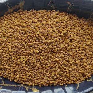 May include: A close-up of a dark bowl filled with dried fenugreek seeds. The seeds are small, irregularly shaped, and light brown. The bowl has a black and gold rim. The seeds are a common ingredient in Indian cuisine.