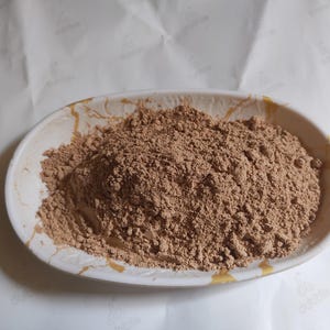 Könnte beinhalten: Eine weiße ovale Schale mit braunem Pulver darin. Das Pulver ist hellbraun und hat eine leicht körnige Textur.