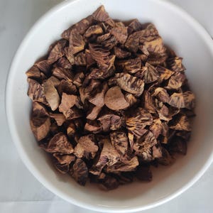 May include: A white bowl filled with chopped, brown betel nuts. The nuts have a textured appearance and vary in size. The bowl is set against a white background, providing a clean contrast to the dark nuts.