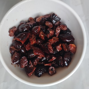 May include: A white bowl filled with dark red, glistening pieces of fruit. The fruit appears to be cut into segments, with some showing the inner flesh. The bowl is set against a white background.