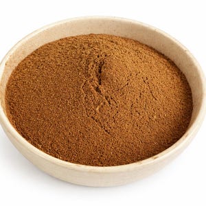 以下が含まれることがあります： ベージュ色の陶器のボウルに茶色の粉末が入っています。粉末はきめが細かく、粒状の質感で、豊かでアースカラーです。丸いボウルはわずかに広がった縁があり、白い背景に配置されています。