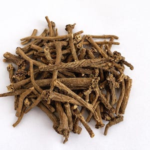 Pode incluir: Um monte de pedaços de raiz de valeriana seca, castanhos, sobre um fundo branco. As raízes têm uma aparência áspera e texturizada com comprimentos e espessuras variáveis. A imagem é um close-up, destacando os detalhes do produto herbal.