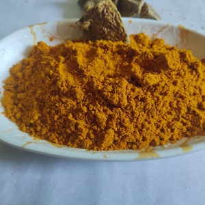 Puede incluir: Un plato de cerámica blanco lleno de polvo de cúrcuma amarillo brillante. Se puede ver un trozo de raíz de cúrcuma seca en el fondo.