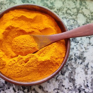 Pode incluir: Uma tigela de madeira cheia de pó de açafrão amarelo brilhante. Uma colher de madeira está apoiada no pó.