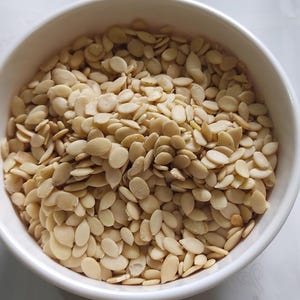 May include: A white ceramic bowl filled with a large quantity of light tan, oval-shaped seeds. The seeds are densely packed, filling the bowl to the top. The bowl is set against a white background.