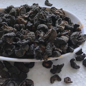 May include: A white ceramic dish filled with dried, dark brown, wrinkled fruit pieces. The fruit is scattered on a white surface.