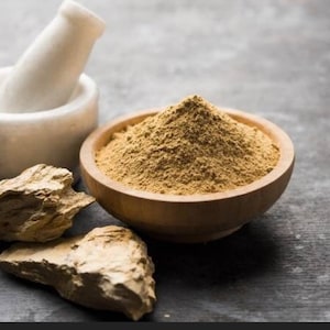 May include: A wooden bowl filled with a light brown powder, next to two pieces of rough, light brown rock. A white mortar and pestle are also in the image.