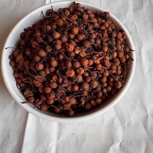 May include: A white bowl filled with dried, round, reddish-brown berries. The berries have small stems and are densely packed. The bowl sits on a white, textured surface, creating a contrast with the dark berries.