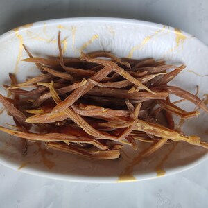 May include: A white oval-shaped bowl with a gold marble design holds a pile of dried, brown, and yellow edible plant stems. The stems are long, thin, and have a slightly curved shape. The bowl is on a white surface.