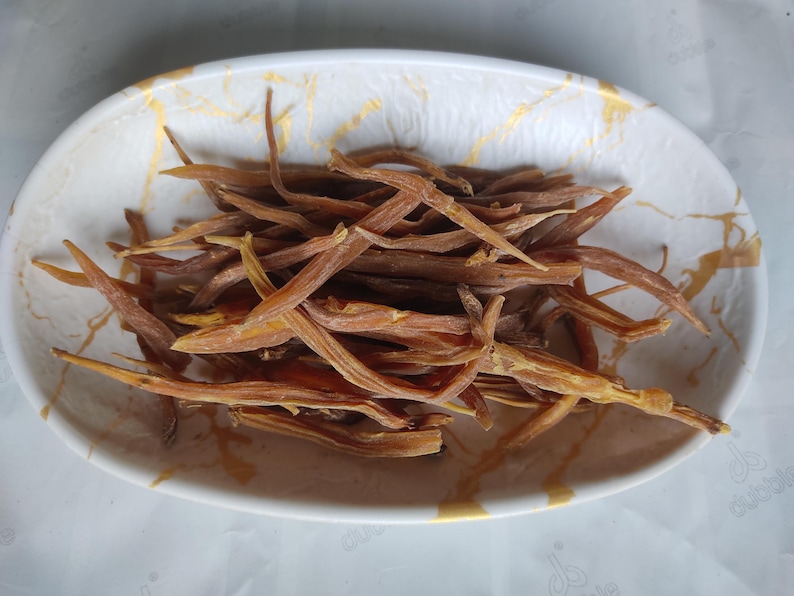 May include: A white oval-shaped bowl with a gold marble design holds a pile of dried, brown, and yellow edible plant stems. The stems are long, thin, and have a slightly curved shape. The bowl is on a white surface.