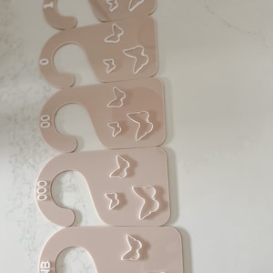 May include: A set of five beige acrylic clothing size dividers. Each divider has a rounded hook shape and a rectangular base. White butterfly outlines decorate the base. Sizes include 1, 0, 00, 000, and NB.