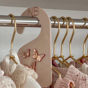 May include: A pink clothing hanger with the letters "NB" in rose gold. The hanger features three rose gold butterfly accents. Several gold-colored hangers hold pink and white clothing.