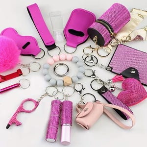 Puede incluir: Una colección de llaveros y accesorios rosas y de pedrería, que incluyen un pompón esponjoso, un estuche de pintalabios, una pulsera, un llavero en forma de corazón y un bolso pequeño. Otros artículos incluyen un soporte para botellas, un abrebotellas y un frasco de desinfectante de manos.