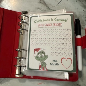 May include: A red binder with a savings tracker sheet that reads "Christmas is Coming!" and "$1000 Savings Tracker." The tracker features a Grinch illustration and heart shapes to fill in. A black marker with a red cap is visible.