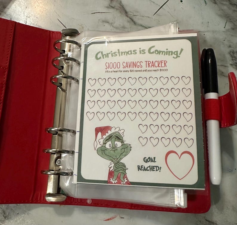 May include: A red binder with a savings tracker sheet that reads "Christmas is Coming!" and "$1000 Savings Tracker." The tracker features a Grinch illustration and heart shapes to fill in. A black marker with a red cap is visible.