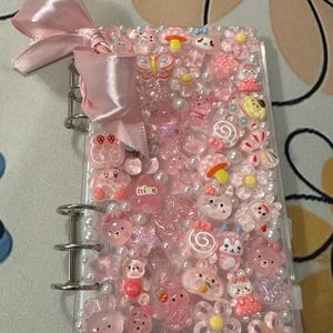 May include: A clear binder decorated with pink embellishments, including flowers, bears, butterflies, and pearls. A pink satin bow adorns the top. The binder is filled with various pink and clear charms and beads.