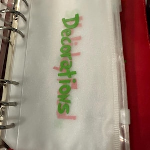 May include: A clear plastic binder pouch with the word "Decorations" in green and pink. The pouch is zippered and attached to a silver binder ring. The binder is red.