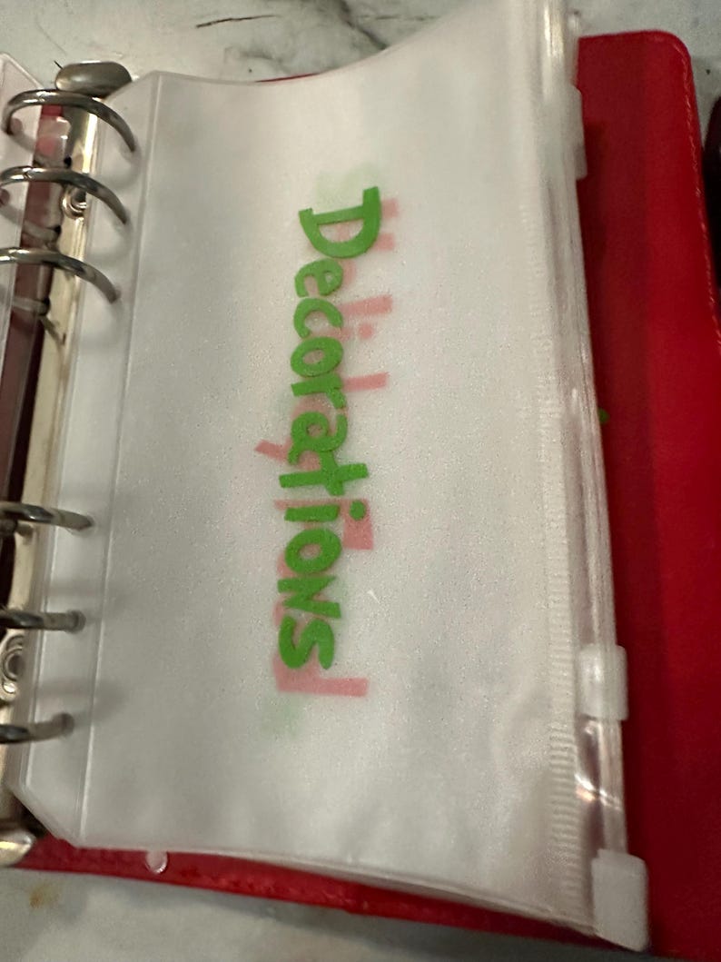 May include: A clear plastic binder pouch with the word "Decorations" in green and pink. The pouch is zippered and attached to a silver binder ring. The binder is red.