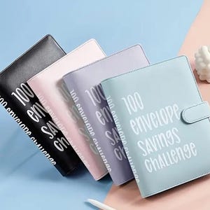 May include: Four faux leather binders in black, pink, lavender, and light blue, for the 100 envelope savings challenge. Each binder has the text "100 envelope savings challenge" printed in white.