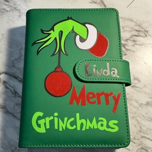 May include: Green wallet featuring a Grinch-themed design with the Grinch's hand holding a red ornament. The words "Merry Grinchmas" are in green and red, with the name "Linda" on the flap.