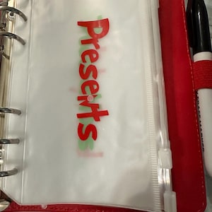 May include: A red binder with silver rings, holding a clear plastic pouch with the word "Presents" in red. A black marker with a red band is visible on the right side.
