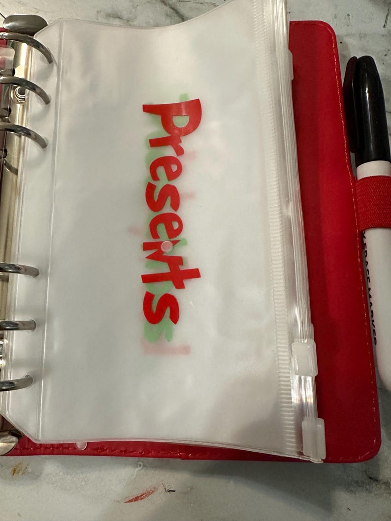 May include: A red binder with silver rings, holding a clear plastic pouch with the word "Presents" in red. A black marker with a red band is visible on the right side.