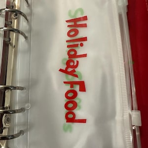 May include: A clear plastic binder pocket with the words "Holiday Food" in red and green lettering. The pocket is inside a red binder with silver metal rings.
