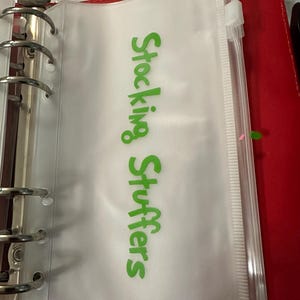 May include: A clear plastic zippered pouch with the words "Stocking Stuffers" in green lettering. The pouch is inside a red binder with silver rings.