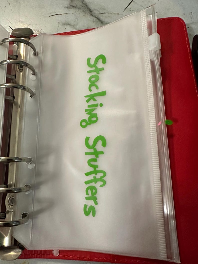 May include: A clear plastic zippered pouch with the words "Stocking Stuffers" in green lettering. The pouch is inside a red binder with silver rings.