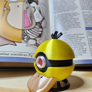 May include: A yellow and black Psyduck figure with a tan beak and red eye, set against an open book with anatomical illustrations. The figure has a spherical head and a duck-like bill. The book's text is partially visible.