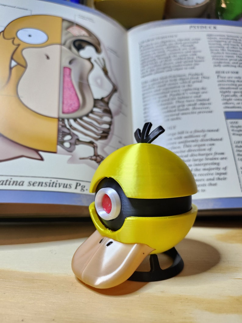 May include: A yellow and black Psyduck figure with a tan beak and red eye, set against an open book with anatomical illustrations. The figure has a spherical head and a duck-like bill. The book's text is partially visible.