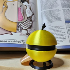 May include: A yellow and black 3D-printed sculpture of a Psyduck character, with a black base and a small beak. The sculpture is in front of an open book with illustrations and text. The book is open to a page with the word "PSYDEER".