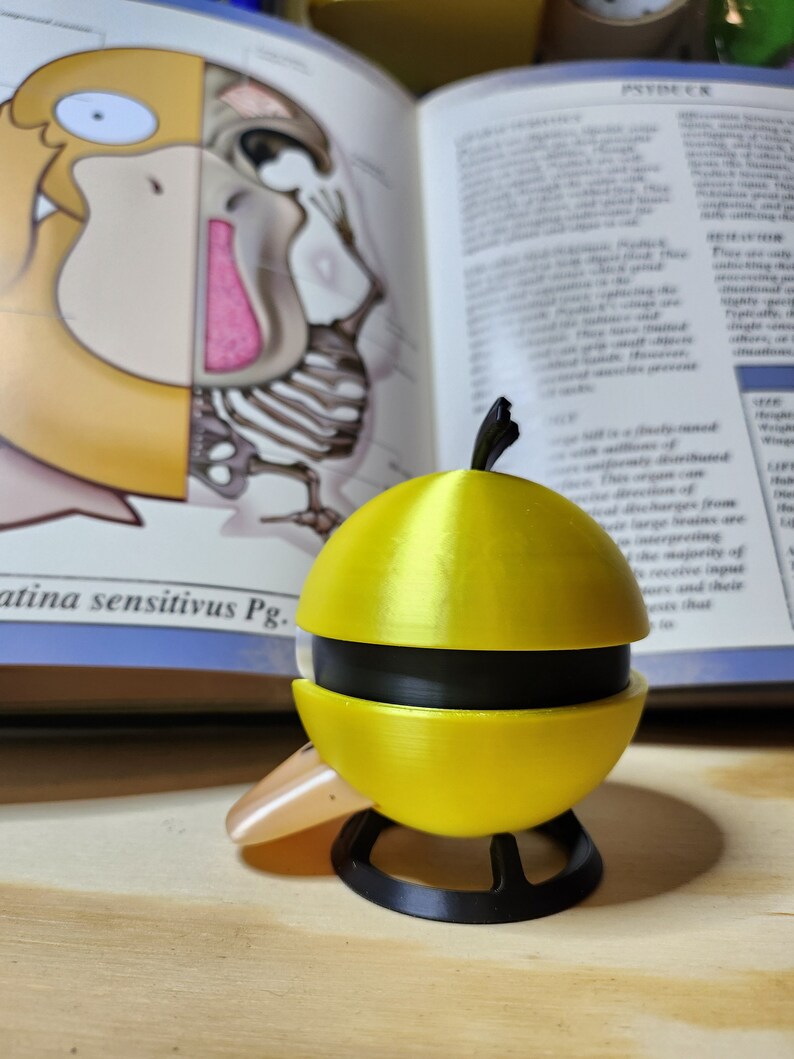 May include: A yellow and black 3D-printed sculpture of a Psyduck character, with a black base and a small beak. The sculpture is in front of an open book with illustrations and text. The book is open to a page with the word "PSYDEER".