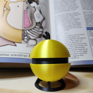 May include: A yellow and black decorative object resembling a stylized fruit, possibly a lemon or a pear. The object is segmented and sits on a black stand. The background includes an open book with illustrations and text.