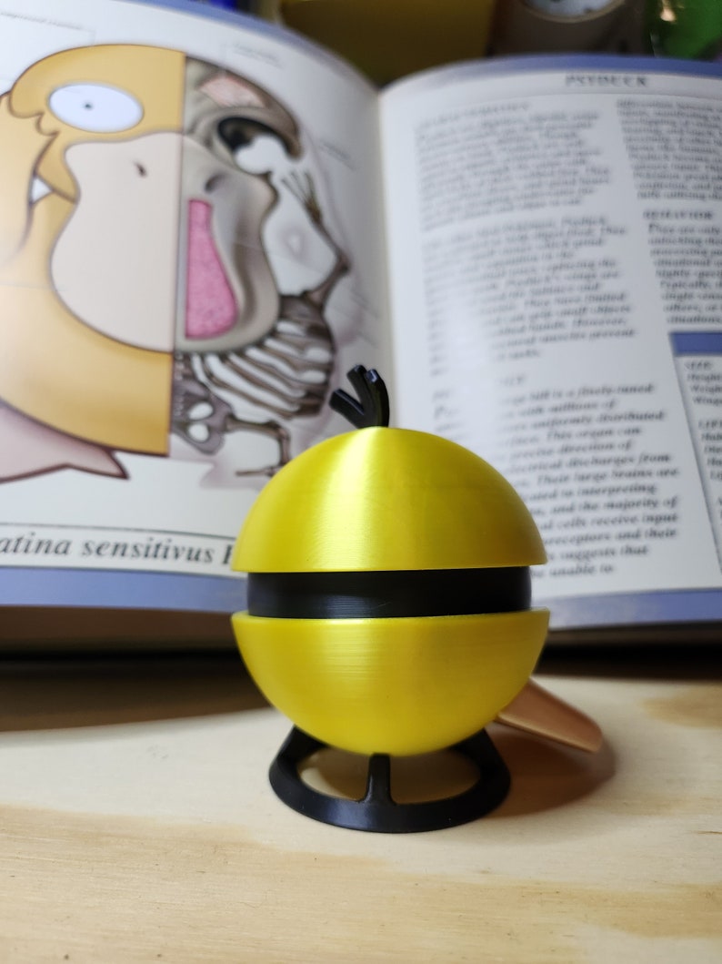 May include: A yellow and black decorative object resembling a stylized fruit, possibly a lemon or a pear. The object is segmented and sits on a black stand. The background includes an open book with illustrations and text.