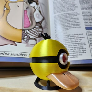 May include: A yellow and black Psyduck-themed 3D-printed model with a duck bill and a red eye. The model is on a wooden surface, with a book open in the background. The book has text and illustrations.
