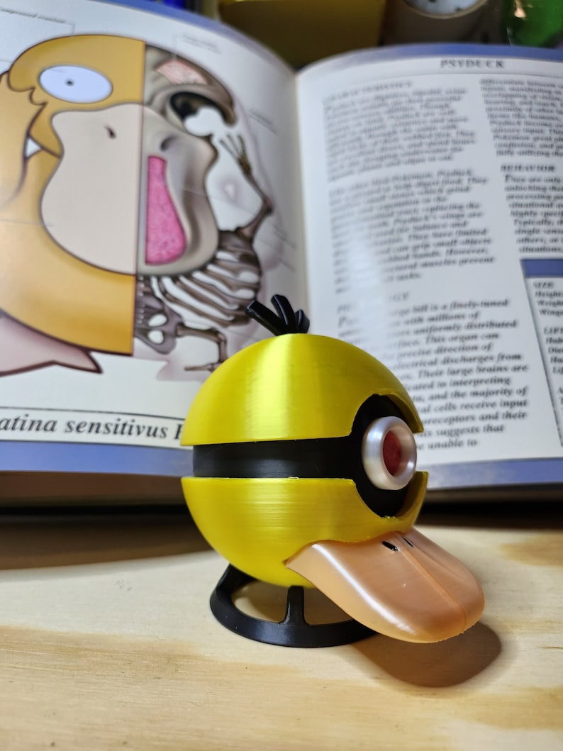 May include: A yellow and black Psyduck-themed 3D-printed model with a duck bill and a red eye. The model is on a wooden surface, with a book open in the background. The book has text and illustrations.