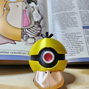 May include: A yellow and black Psyduck figure with a red eye, based on the Pok&eacute;mon character. The figure is 3D-printed and has a duck-like bill. The background includes an open book with anatomical illustrations and the word "PSYDUCK".
