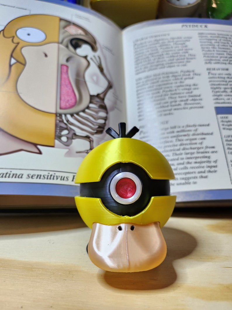 May include: A yellow and black Psyduck figure with a red eye, based on the Pok&eacute;mon character. The figure is 3D-printed and has a duck-like bill. The background includes an open book with anatomical illustrations and the word "PSYDUCK".