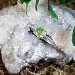Könnte beinhalten: Ein silberner Ring mit einem grünen Peridot-Edelstein. Der Ring liegt auf einem Bett aus weißen Kristallen.