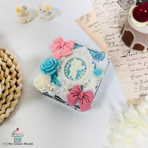 Puede incluir: Una caja cuadrada decorativa adornada con lazos rosas, rosas azules y blancas y un camafeo. La caja presenta un fondo azul claro con encaje blanco y detalles de perlas. Dos pequeñas figuras de gato están en el fondo.