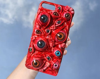Puede incluir: Una funda de teléfono roja con un diseño de varios ojos con pupilas de diferentes colores. Los ojos están sobre un fondo rojo y parecen sobresalir de la funda.