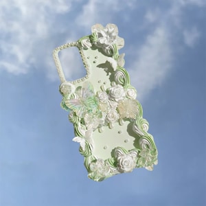 May include: A green and white phone case with a 3D design featuring flowers, butterflies, and pearls. The case has a raised, textured design and a cutout for the phone's camera.