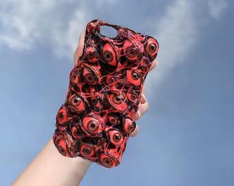 Puede incluir: Una funda de teléfono roja y negra con un diseño de muchos ojos. Los ojos son de diferentes tamaños y formas, y están dispuestos en un patrón aleatorio. La funda está hecha de un material suave y flexible.
