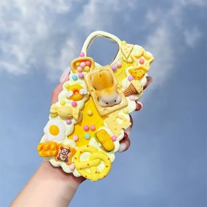 May include: A yellow phone case decorated with various food items, including a fried egg, a pretzel, a hamburger, a bear, and a piece of cheese. The case is made of resin and has a 3D effect.
