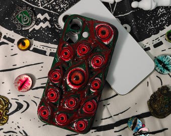 Puede incluir: Una funda de teléfono verde y roja con un patrón de múltiples globos oculares rojos. La funda está junto a un teléfono blanco y varias piezas decorativas de vidrio con forma de ojo. La funda tiene un acabado brillante.